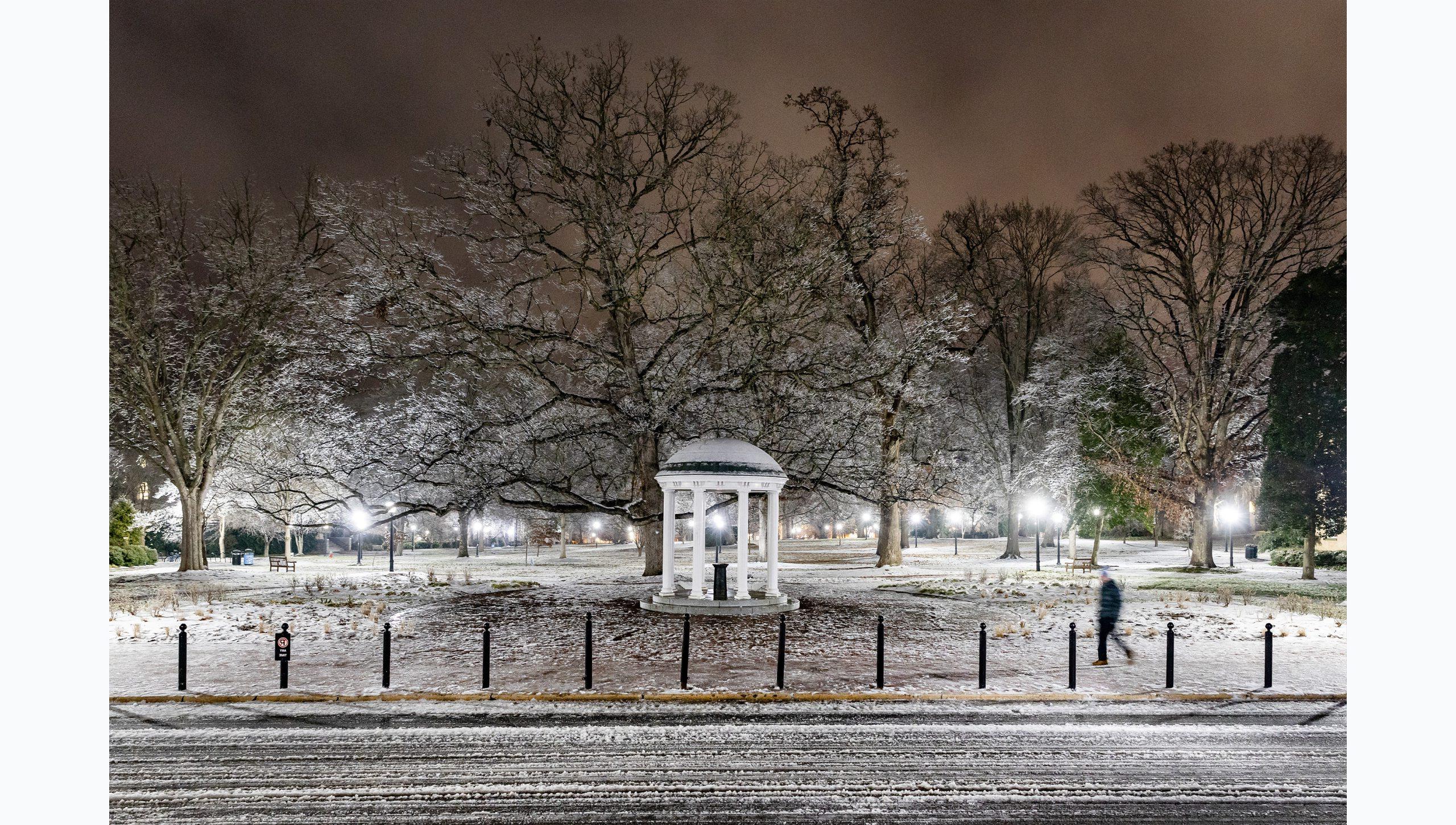 Pre-dawn photo of the Old Well covered in snow and ice on the campus of U.N.C Chapel Hill. One person is seen walking in front of the well, and McCorkle Place is seen in the background.